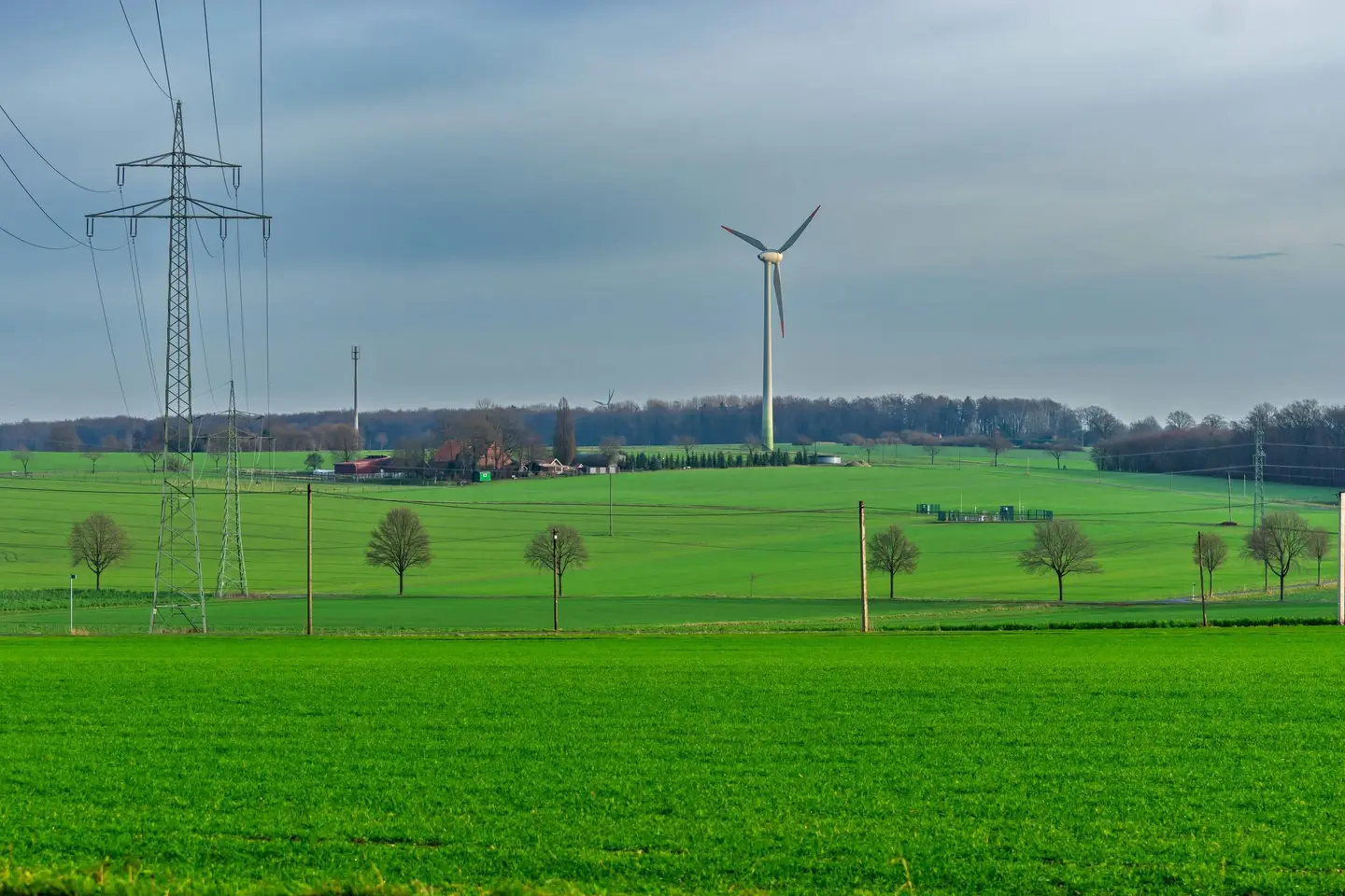 A wind turbine and power lines standing over green fields under a gray sky.